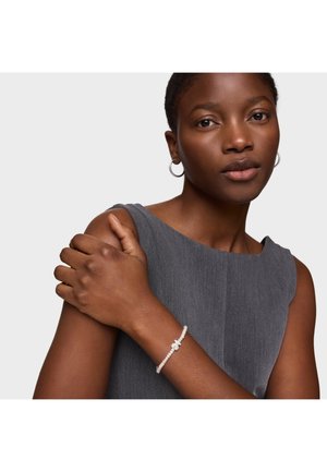 Młoda kobieta z krótkimi włosami, nosząca kolczyki koła, szary top bez rękawów i koralikową bransoletkę, trzymająca się za przeciwne ramię.