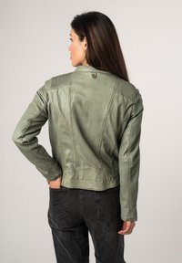 Giacca leggera verde oliva con finitura testurizzata, design aderente e cuciture pronunciate sulle spalle, abbinata a jeans neri.