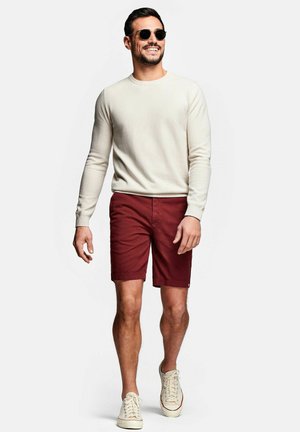 Homme souriant portant des lunettes de soleil rondes, une chemise crème à manches longues, un short rouge et des baskets blanches marchant devant un fond clair uni.