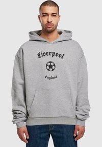 Grå hettegenser med lomme foran, med svart tekst som sier "Liverpool" og et fotballgrafikk. Uformell design, myk stoff.