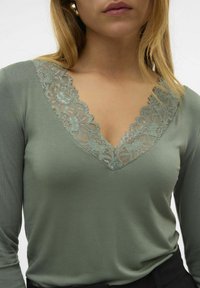 Top de manga larga en tela verde suave con un delicado escote en V de encaje con patrones florales, textura suave y diseño ajustado.