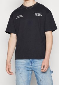 Camiseta de algodón negra de manga corta con texto blanco "LOS FUMADORES MUEREN JOVEN" y "PEQUS", ajuste relajado, cuello redondo y textura suave.