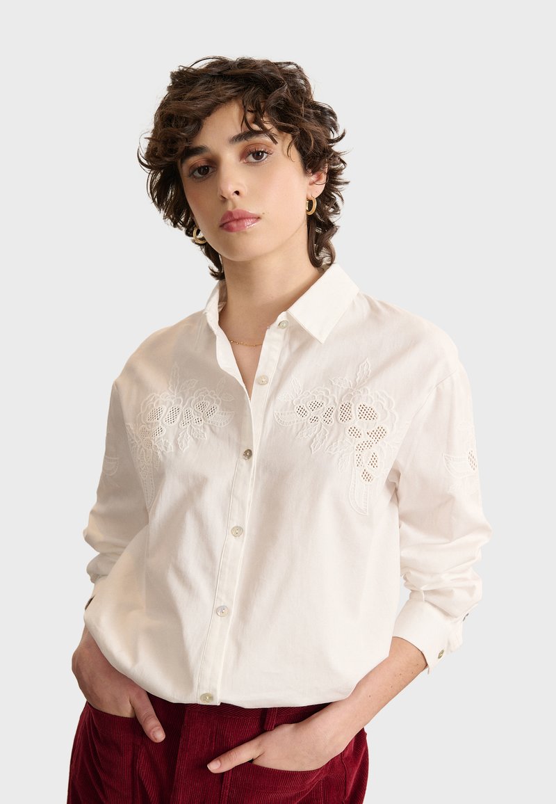 Camicia bianca a maniche lunghe con ricami floreali sulle spalle, sul colletto e sulla parte anteriore con bottoni. Tessuto in cotone morbido. Abbinata a pantaloni in velluto a coste rossi.