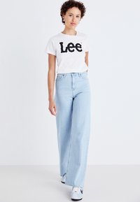 Vit t-shirt med svart "Lee" logotyp, högmidjade ljusblå vida jeans och vita sneakers med svarta detaljer. Slät tyg, avslappnad passform.