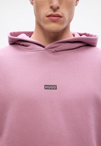 Tricou cu glugă în roz deschis, realizat dintr-un material texturat. Prezintă un logo negru "HUGO" pe piept. Design simplu și minimalist.