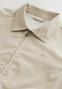 Chemise zippée beige avec un col, présentant une texture subtile, une doublure à rayures contrastantes et un petit logo brodé sur la poitrine.
