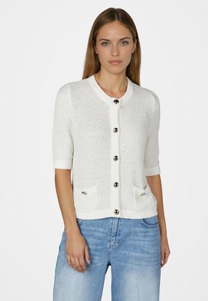 Frau trägt eine weiße, strukturierte Kurzarm-Strickjacke mit silbernen Knöpfen und Fronttaschen, kombiniert mit hellblauen Jeans, stehend vor weißem Hintergrund.