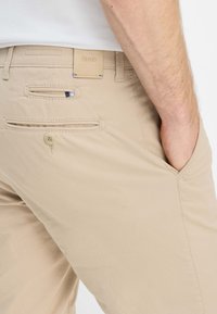 Beige broek met een gladde textuur, voorzien van achterzakken en een leren merkembleem. Zijkant zak zichtbaar, met subtiele stiksel-details.