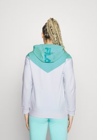 Sudadera de color púrpura claro con capucha y panel trasero en color turquesa. Presenta un diseño en forma de V con un logo en la parte posterior. Textura suave, corte casual.