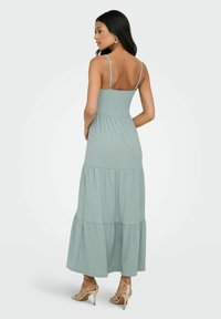 Robe maxi vert menthe avec un corsage smocké, des bretelles fines et une jupe à volants. Le tissu est doux avec une texture légère et fluide.