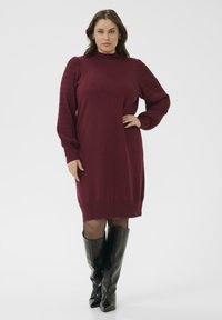 Burgunderfarbendes Strickkleid mit gerippten Ballonärmeln, Rundhalsausschnitt und lockerer Passform, kombiniert mit kniehohen schwarzen Stiefeln.