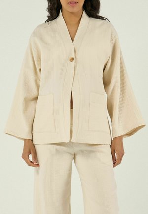 Vrouw die een crèmekleurige twee-delige outfit draagt met een gestructureerde stof, wijde mouwen, voorzakken en een enkele bruine knoop op het bovenstuk.