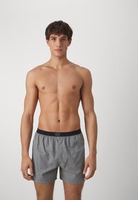 Šedé vzorované boxerky s černým elastickým pasem, vyrobené z lehkého materiálu, prezentované na modelovi bez trička na neutrálním pozadí.