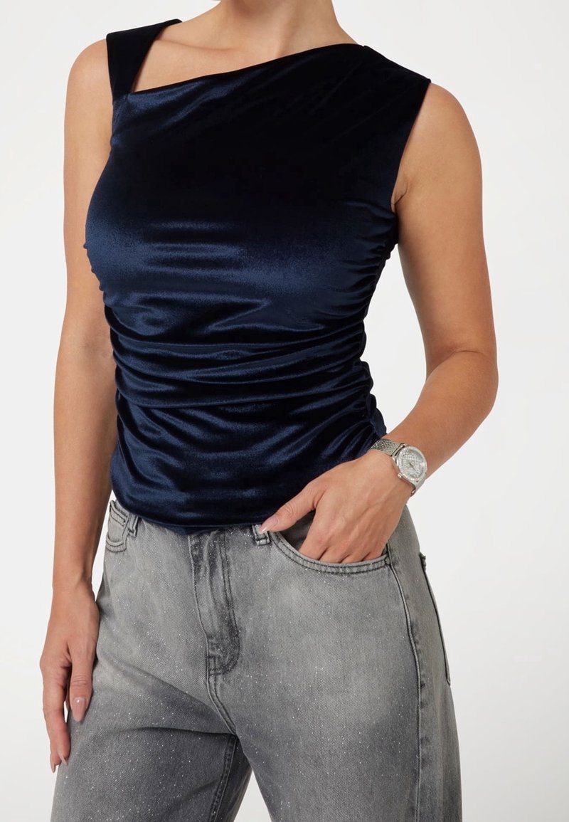 Femme portant un haut sans manches en velours bleu marine et un jean gris tacheté, une main dans la poche et une montre argentée au poignet.