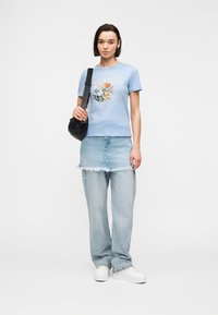 Femme portant un t-shirt graphique floral bleu clair, une jupe en jean superposée sur un jean, des baskets blanches, et portant un sac à bandoulière noir.