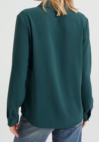 Mörkgrön långärmad skjorta med en slät textur, med krage och knäppta cuffs, sedd bakifrån med en oval fåll.