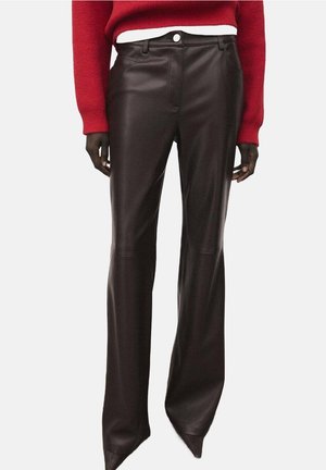 Person trägt eine dunkelbraune Lederhose mit hohem Bund, kombiniert mit einem roten Pullover und einem weißen Hemd darunter, steht vor einem schlichten Hintergrund.