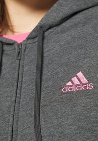 Sweat à capuche gris zippé en tissu lisse, avec un logo Adidas rose brodé sur le côté gauche de la poitrine et un cordon de serrage noir.