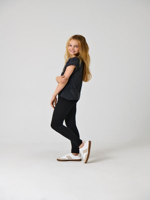 Sort kortærmet T-shirt, sorte leggings og hvide sneakers med brune såler. Lige hår, smilende, stående i en afslappet positur.