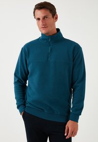 Türkiser Fleece-Pullover mit hohem Kragen, Viertelreißverschluss, langen Ärmeln und gerippten Bündchen. Verfügt über eine horizontale Naht über der Brust.