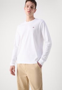 Camisa blanca de manga larga de algodón con un pequeño logo en el pecho, combinada con pantalones caqui. Diseño sencillo y textura suave.