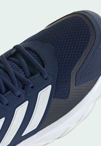 Gros plan sur une chaussure de sport bleu marine en tissu mesh, avec des lacets épais, une semelle blanche et des bandes réfléchissantes blanches sur le côté.