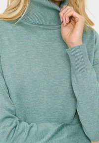 Pull à col roulé en tricot vert menthe, avec poignets et col côtelés. Texture douce avec une coupe ajustée, mettant en valeur la chaleur et le confort.