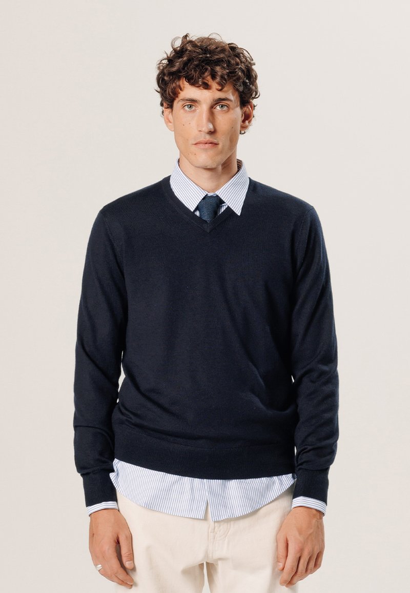 Pull marin en V en tissu tricoté doux, porté sur une chemise à rayures bleu clair, avec des poignets visibles et une coupe décontractée.