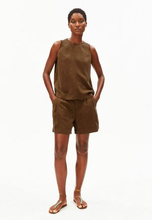 Vrouw staand met handen in de zakken, draagt een bruin mouwloos topje, bijpassende korte broek en bruine sandalen met enkelbandjes op een witte achtergrond.