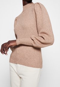 Suéter beige claro acanalado con cuello alto y mangas abullonadas; hecho de tela suave y texturizada; combinado con pantalones de color crema.