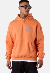 Sweat à capuche orange avec une poche dans le devant, un cordon de serrage pour la capuche et un logo brodé. Le tissu semble doux, avec des poignets et un ourlet côtelés.