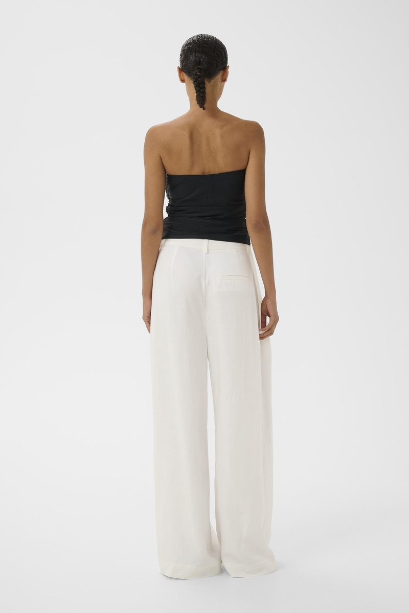 Frau mit geflochtenem Haar steht mit dem Rücken zur Kamera, trägt ein schwarzes, trägerloses Oberteil und weite, weiße Hosen vor einfarbigem Hintergrund.