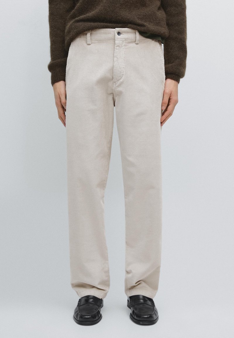 Personne portant un pantalon en velours côtelé beige, un pull marron foncé et des mocassins noirs en cuir, debout devant un fond gris clair.