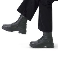 Zwarte leren legervoetbalschoenen met een gestructureerde afwerking, vetersluiting en stevige rubberen zolen. De broek is zwart en cropped net boven de enkel.