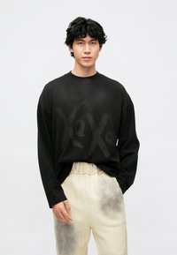 Melnas lielizmēra džemperis ar izbalējušu logotipa dizainu. Garas piedurknes, apaļa izgriezuma kakls, valkāts kopā ar gaiši bēšām sporta biksēm ar pelēkiem gradientiem.