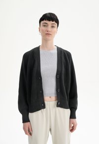 Schwarze Strickjacke mit gerippten Bündchen und Saum, mit Knopfverschluss, getragen über einem hellgrauen, kurzen Oberteil und hellen Hosen.