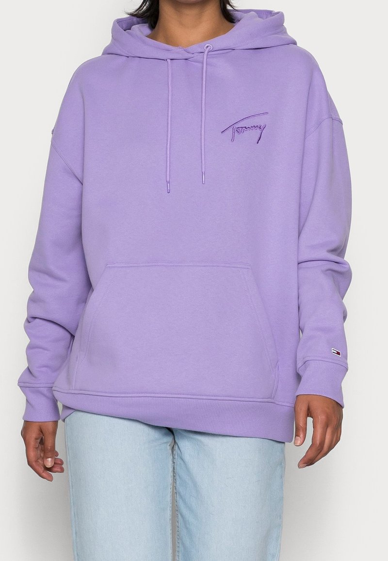 Personne portant un sweat à capuche lavande ample avec poche avant et logo brodé "Tommy", associé à un jean bleu clair.
