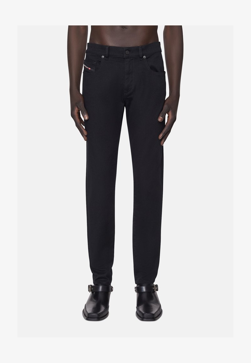 Fekete farmernadrág, slim fit szabással, elülső zsebekkel, egy kis logóval a derékpánt közelében, és megerősített varrásokkal. Fekete cipővel viselve.