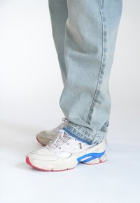 Zapatillas deportivas gris claro con acentos en rojo, azul y blanco, combinadas con unos vaqueros sueltos de color azul claro con un dobladillo desgastado y un patrón sutil.