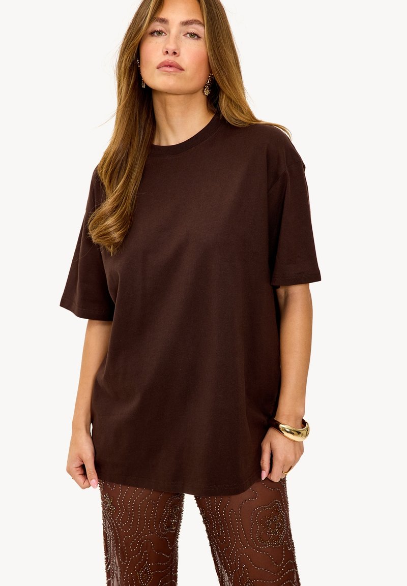 T-shirt oversize marron en coton doux, avec un col rond et des manches courtes, associé à un pantalon en tissu transparent à motifs intricats.