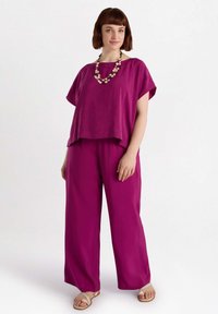 Fuksjowa lniana bluzka z krótkim rękawem, zestawiona z pasującymi spodniami o szerokich nogawkach. Wyrazisty chunky naszyjnik i płaskie sandały dopełniają stylizację.