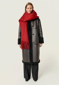 Lang sort shearling frakke med en blank finish, sorte manchetter og et rødt frynset tørklæde. Sammen med brede sorte bukser.