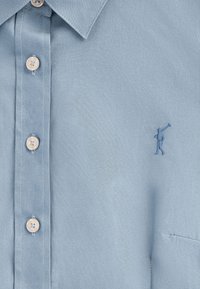 Camicia button-up azzurro chiaro realizzata in tessuto morbido, con una sottile figura ricamata sul lato sinistro e bottoni beige.