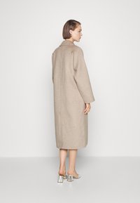 Beige wollen jas met een losse pasvorm, tot halverwege de kuit, en lange mouwen. Kenmerkt zich door een rechte rugnaad en een seliekragen. Gedirigeerd met zilveren hakken.