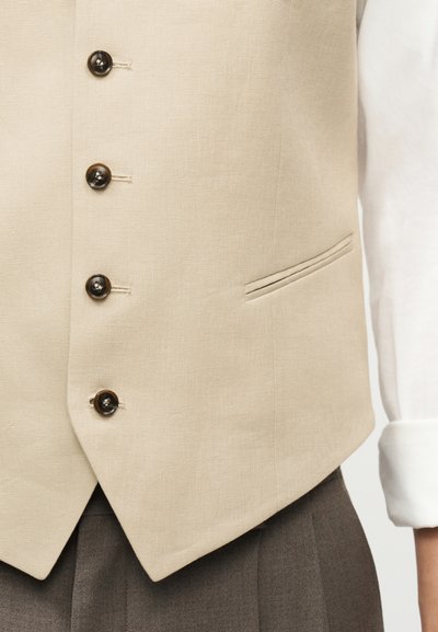 Μπεζ γιλέκο με κουμπιά και πλαϊνή τσέπη, φορεμένο πάνω από λευκό πουκάμισο με μακριά μανίκια και συνδυασμένο με γκρι παντελόνι.