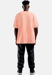 T-shirt rosa chiaro a maniche corte con vestibilità ampia, abbinata a pantaloni neri e sneakers, vista da dietro. Design semplice, senza motivi visibili.