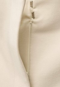Hellbeige Stoff mit Struktur, versehen mit einem Reißverschluss und zwei Metallverschlüssen entlang der Naht, was auf ein strukturiertes Design hinweist.
