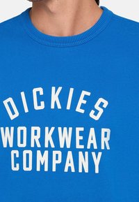 Modrá bavlněná mikina s žebrovaným kulatým výstřihem a velkým bílým nápisem "DICKIES WORKWEAR COMPANY" na přední straně.