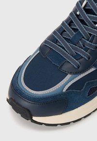 Sneaker v mornarsko modri barvi, ki vključuje mešane materiale, gladko usnje, teksturiran mrežast material, odsevne detajle in gumijast podplat.