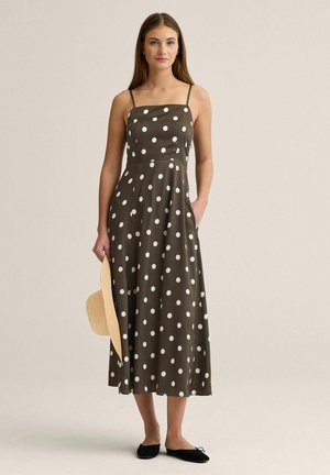 Kvinde iført en ærmeløs brun midikjole med hvide polkaprikker, der holder en stråhat og har sorte flade sko på, stående foran en enkel baggrund.